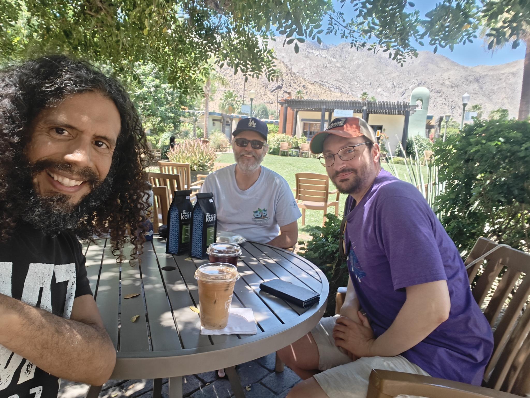 Three friends sitting outside enjoying coffee and conversation. In the background are brown mountains